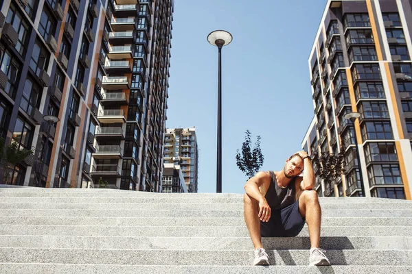 Un giovanotto che si allena fuori. Stanco atleta esausto seduto da solo su gradini esterni vista urbana e riposo dopo runing o duro allenamento. Abbi cura di te. Sedetevi in posizione accovacciata. — Foto Stock