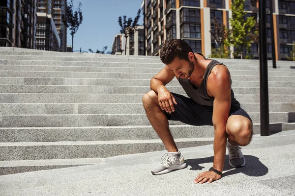 Genç adam dışarıda egzersiz yapıyor. Ciddi bir adam asfalta dokunuyor ve aşağıya bakıyor. Egzersiz, koşu ya da koşudan sonra dinlenin. Basamakların yanında tek başına otur..