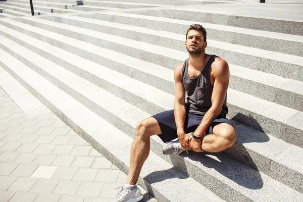 Young man exercising outside. Hot good-looking sexy handsome athlete posing on camera by sitting on white steps outside. Hold his leg with hands and look straight on camera.