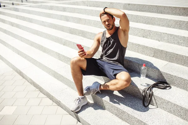 Young man exercising outside. Sexy hot athlete sitting on white steps outside and posing on camera. Guy hold phone in hand. Rubber band and water bottle on steps besides. Rest after workout.