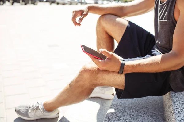 Genç adam dışarıda egzersiz yapıyor. Elinde telefon tutan sporcunun yan görüntüsünü kes. Bilek çevresindeki spor takip cihazı. Antrenmandan sonra dinlenin. Modern teknoloji ve aygıtlar.