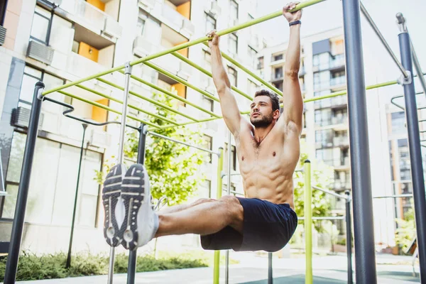 Young man exercising outside. Strong powerful well built guy hang on the horizontal bar and holding legs in horizontal position towards ground. Training body. — Stock Image Young man exercising outside. Strong powerful well built guy hang on the horizontal bar and holding legs in horizontal position towards ground. Training body. Royalty Free Stock Images
