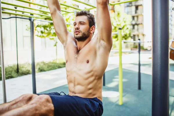 Young man exercising outside. Close up and cut view of strong powerful well built muscled guy hang on the horizontal bar. Looking forward. Legs are in horizontal position towards ground. — Stock Image Young man exercising outside. Close up and cut view of strong powerful well built muscled guy hang on the horizontal bar. Looking forward. Legs are in horizontal position towards ground. Royalty Free Stock Photos