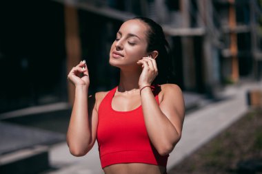 Kırmızı spor tişörtlü, siyah atkuyruklu, kulaklık takan ve müzik dinleyen rahat genç bir kadın. Şehir sporu konsepti. Arka planda mavi apartman bloğu.