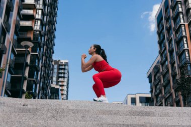 Siyah at kuyruklu, merdivenlerin üstüne çömelmiş atletik güzel bir kız. Sportif kadın dışarıda bacak kaslarını çalıştırıyor. Arkaplanda apartman blokları.