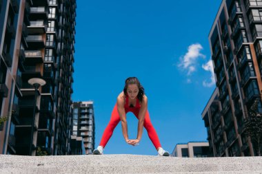 Kırmızı spor giyim giymiş, forvet ve esneme hareketi yapan çekici genç bir kadının ön görüntüsü. Kırmızı spor sutyen ve tozluklu kız. Düşük açılı çekim. Arkaplanda apartman blokları.