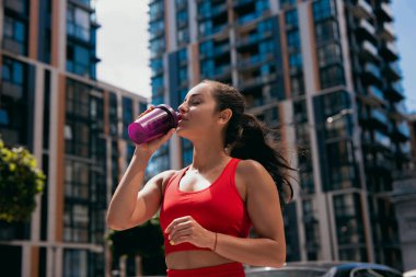Koşudan sonra spor şişesinden su içen kırmızı sütyenli genç bir kadının portresi. Kahverengi dalgalı saçlı sportif kız. Elindeki kırmızı bilezik. Arka planda bulanık apartman blokları.