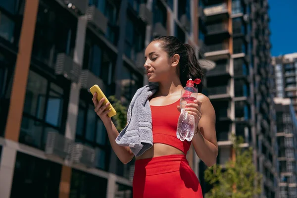 Belle jeune femme sportive aux cheveux bruns portant un haut rouge et des leggins prenant une pause après l'entraînement ou le jogging. Fille regardant smartphone et tenant bouteille en plastique avec de l'eau dans sa main gauche. — Photo