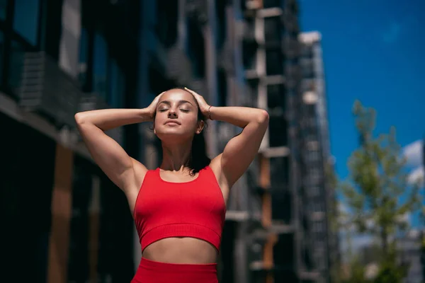 Fit jovem mulher com rabo de cavalo escuro vestindo sportswear vermelho. Menina desportiva com os braços levantados tocando seu cabelo e sorrindo. Fundo turvo da cidade. Apartamento bloco e árvores no fundo. — Fotografia de Stock