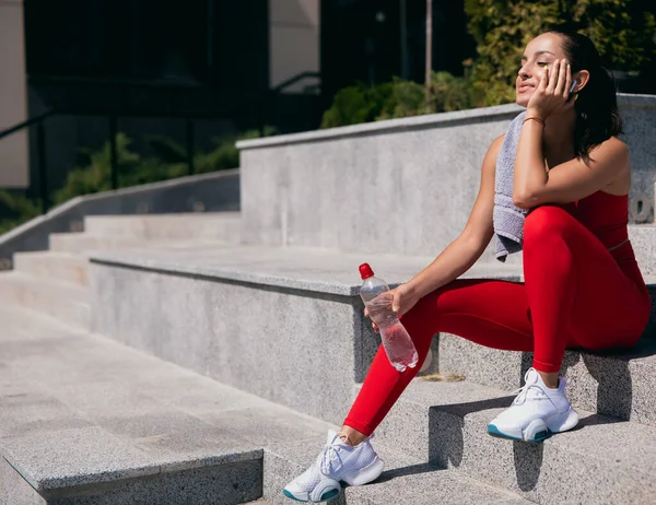 Jovem bem construída em top de esportes vermelhos, leggins e tênis brancos sentados nas escadas, seu rosto apoiado em sua mão. Ela tem uma garrafa de água na mão direita. Violeta toalha pendura em seu ombro. — Fotografia de Stock