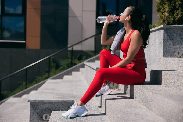 Sidovy över vinden ung kvinna bär röd topp och leggins nd vita sneakers sitter på trappor. Flicka tar paus och dricker vatten från plastflaska. Stadsidrott. — Stockfoto