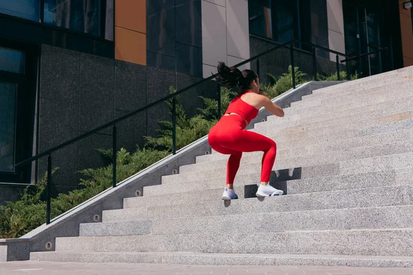 Mulher fitness em sportswear vermelho e tênis branco, com fones de ouvido sem fio pulando em escadas. Treino cardiológico. Bloco de apartamentos no fundo. Treino no conceito de cidade grande. — Fotografia de Stock