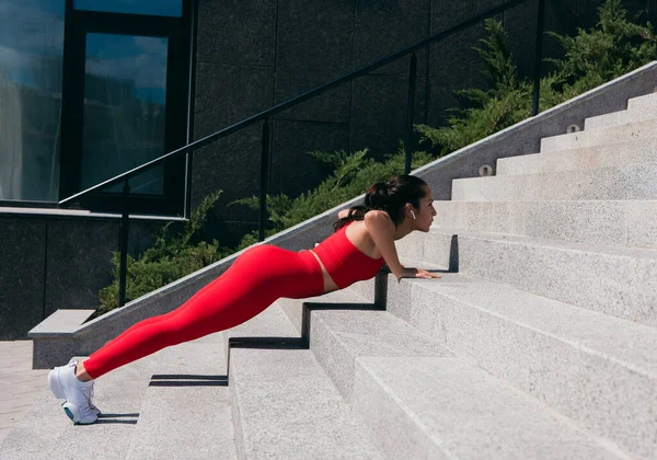 Vista lateral da jovem bem construída empurrando para cima das escadas. Mulher atlética com cabelo castanho vestindo top vermelho e leggins, fones de ouvido brancos e tênis trabalhando na rua. — Fotografia de Stock