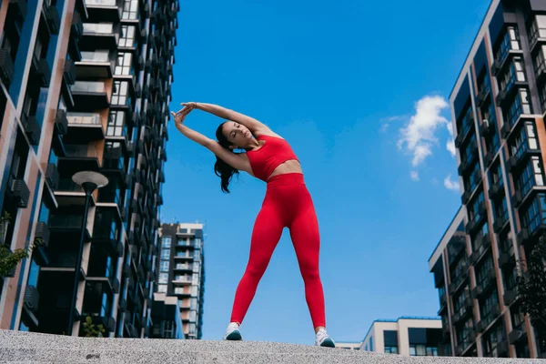 Mulher bonita com cabelo ondulado escuro vestindo top esportivo vermelho e leggins, tênis brancos esticando os braços. Desporto no conceito de cidade grande. Apartamento bloco no fundo. — Fotografia de Stock