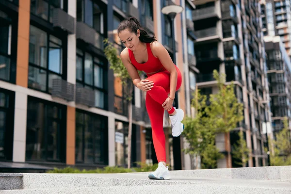 Jovem atraente sentindo dor no pé durante o treino esportivo ao ar livre. Menina em sportswear vermelho e tênis brancos de pé em uma perna e sentindo cãibras em sua perna de lef. Conceito de desporto urbano. — Fotografia de Stock