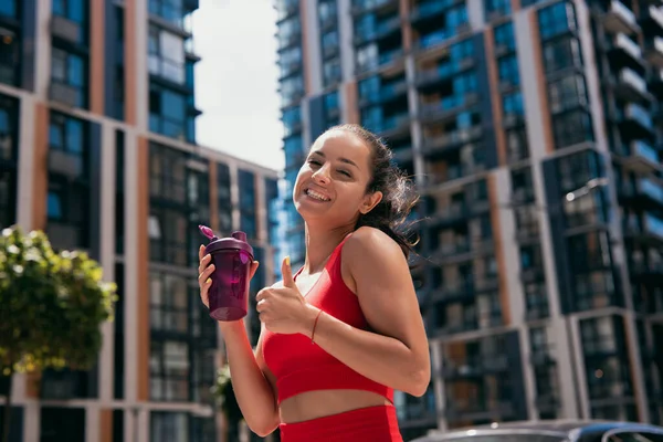 Sporty adorabile sorridente giovane donna con i capelli scuri indossa reggiseno rosso tenendo bottiglia sportiva e pollice in su. Condomini sfocati sullo sfondo. Concetto sportivo urbano. — Foto Stock