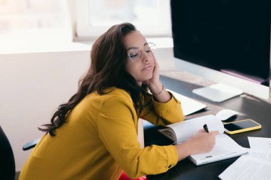 Uyuyan yorgun kadın elinde kırmızı bilezik ile ayakta duruyor. Fazla mesai ve fazla çalışma konsepti. Kahverengi saçlı, yuvarlak gözlüklü kız. Evraklar, bilgisayar ve akıllı telefon masanın üstünde. Evden ya da ofisten çalış..