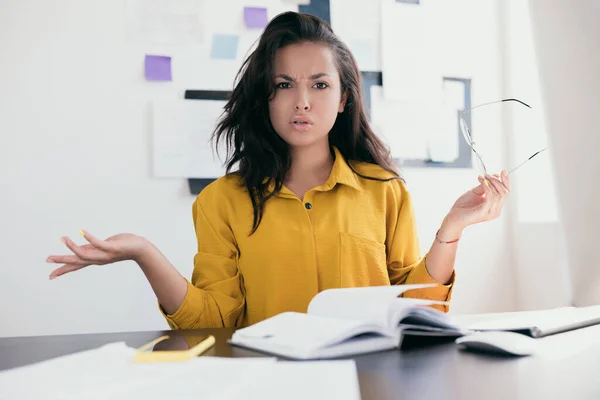 Perturbed office worker. Stressed young woman throw up her hands. Lady in yellow blouse holding glasses in hand. There is an open notebook and phone on the table. Office work work from home concept. — Stock Photo, Image