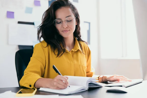 Yoğunlaşmış çekici kadın ofis masasında oturuyor ve defterine planlar yazıyor. Akıllı beyaz kadın ofis çalışanı kağıtları imzalıyor. Ofis işi ya da serbest çalışan konsepti. — Stok fotoğraf