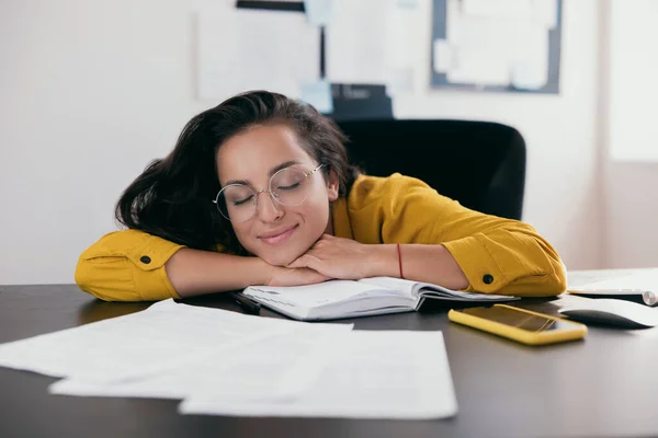 Happy woman with long brown hair wearing bright yellow blouse and round glasses takes break. Her face propped on her hand. Diary, papers and smartphone on desk. Freelance or working at home concept.