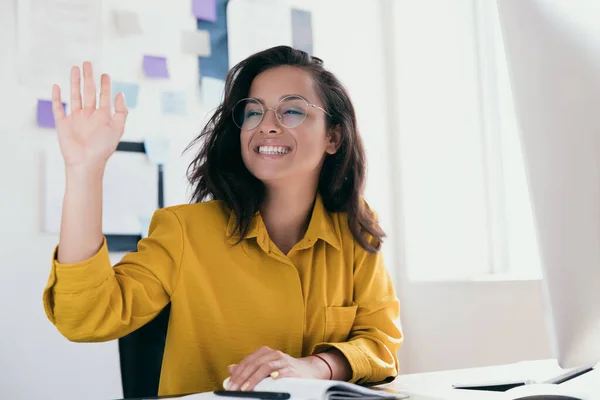 Neşeli genç bayan ofis çalışanı birini selamlamak için elini kaldırdı. Yuvarlak gözlüklü sarı elbiseli kız. Ofis ev konseptiyle çalışıyor ya da çalışıyor. Arkaplanda notlar ve planlar. — Stok fotoğraf
