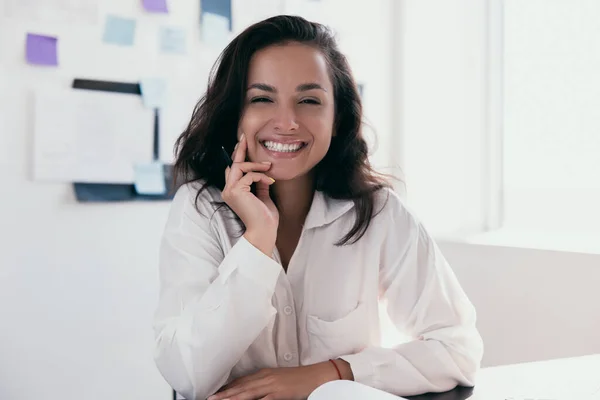 Excited caucasian woman with brown hair sit at desk feeling euphoric and looking at camera. Office or freelance happy female worker. Her face propped on her hand. Freelance or office work concept.
