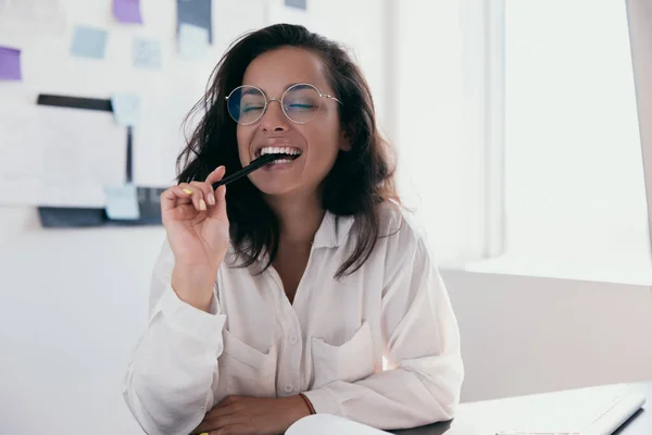 Intelligent businesswoman bite pen with her teeth and smiles happily. Office or freelance female worker in white shirt and round glasses. Positive thinking person concept.