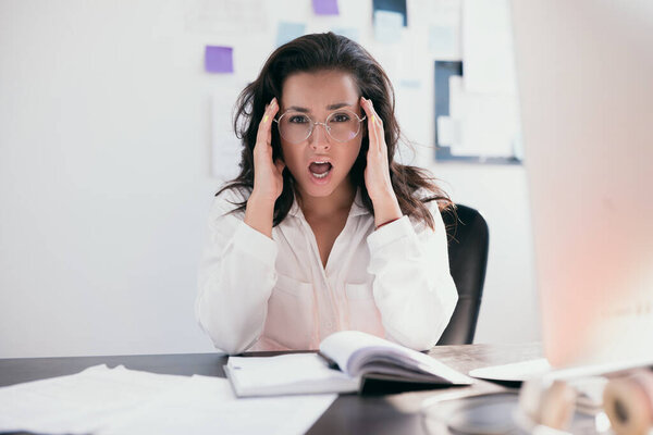 Вид спереди профессиональной молодой женщины с круглыми очками в белой блузке, сидящей у стола с напряженным впечатлением на лице, с открытым ртом. Работник офиса. Фрилансер на рабочем месте.