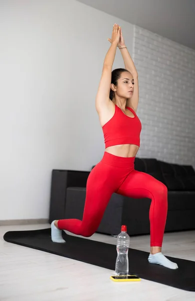 Ung kvinna i röd träningsoverall som tränar eller yogar hemma. Allvarlig, lugn och koncentrerad smal flicka står på ett ben knäböj position och hålla händerna upp tillsammans. Träning och träning. — Stockfoto