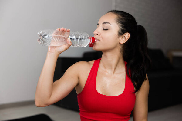 Young woman in red tracksuit doing exercise or yoga at home. Picture or side view of attractive beautiful sporty girl drinking water and hydrating. Sitting alone in room after exercising, training.