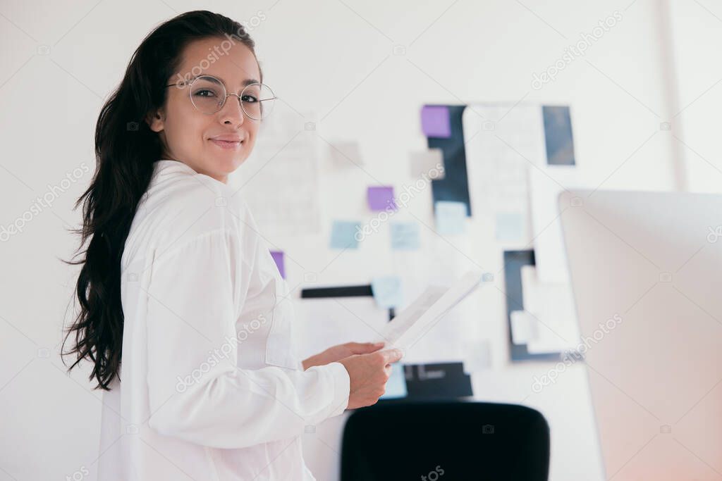 Cheerful beautiful young woman wear white shirt and round shaped glasses which holds papers looking at camera and smiling. Light blurred background. Office work or work at home concept.