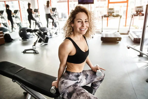 Uma mulher está sentada num aparelho de treino. Ela está olhando para a câmera e sorrindo — Fotografia de Stock