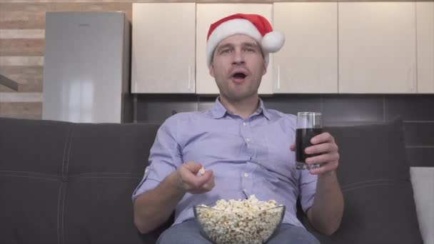 Emotionele feestelijke jongeman in kerstmuts die popcorn eet en frisdrank drinkt tijdens het kijken naar tv, film of film. Meneer alleen op de bank en wissel van kanaal met afstandsbediening. Emotionele gelukkige man. — Stockvideo