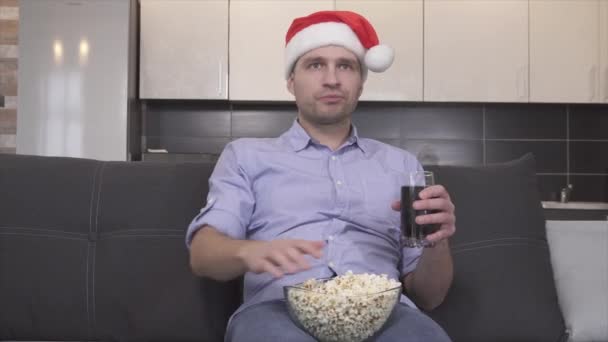 Видео серьезного сконцентрированного парня в рождественской шляпе, который ест попкорн, пьет сиду и смотрит телевизор один в комнате. Переключение каналов с дистанционным управлением. Рождество или Новый год. — стоковое видео