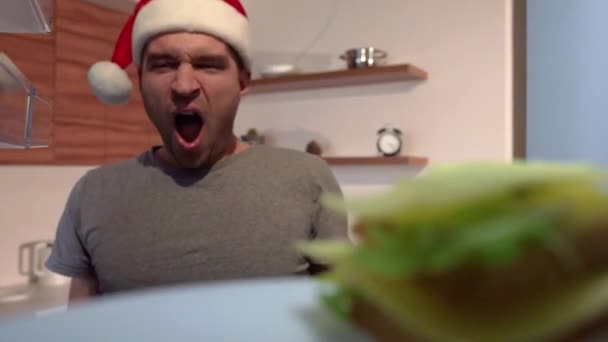 Christmas video of young sleepy man with new year hat openes fridge door and take bitten sandwich to take a bite. Chew snack and put it back on plate and close door. — Stock Video