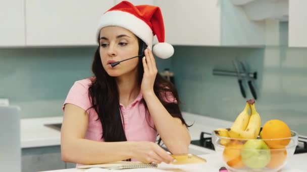 Mujer joven trabajando en casa durante la cuarentena. Tiempo festivo o período de año nuevo. El empleado del centro de llamadas habla en video chat y notas para fumadores. Chica multitarea. — Vídeo de stock