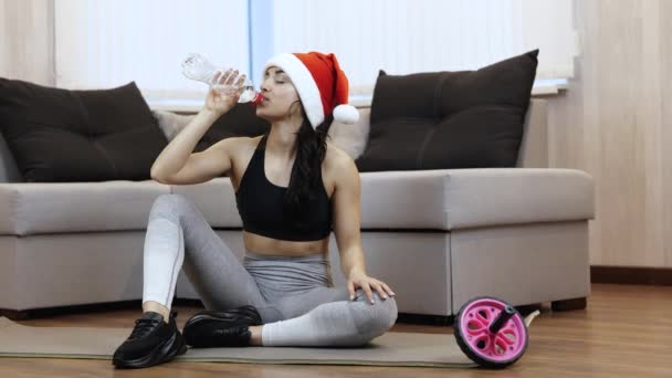 Una mujer joven hace ejercicio en casa durante la cuarentena. Ajuste el resto femenino del modelo y beba el agua de la botella. El tiempo de Navidad o el período de año nuevo. 2021. — Vídeo de stock