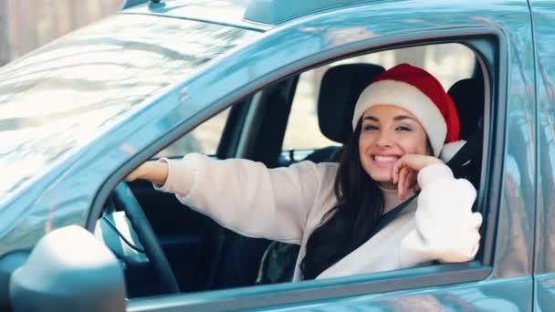 Mujer joven sentada en coche durante la pausa de viaje. Chica positiva feliz usar sombrero de Santa rojo y sonrisa. Levanta los pulgares. Año nuevo o Navidad. Mujer joven es conductor solo en coche. — Vídeos de Stock