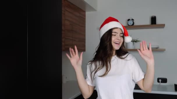 Egy fiatal nő a konyhában a karantén alatt. Karácsonyi kalapos lány táncol és pózol a kamera előtt. Ünneplés új 2021 év örömmel és boldogsággal. — Stock videók