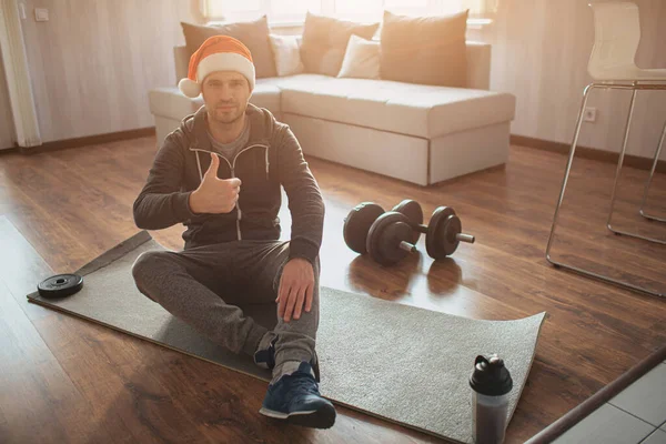 Young ordinary man go in for sport at home. Guy with red hat on his head sit on mat and hold big thumb up. Has rest after first hard wokrout. Winter holdays or Christmas eve.
