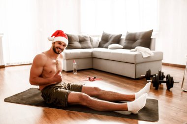 Mutlu Noeller ve Noel Baba 'lı mutlu yıllar kırmızı şapkalı adam evde egzersiz yapıyor. Üstsüz bir halıya oturur ve baş parmağını kaldırır..