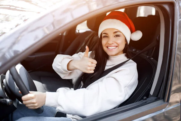 Mutlu yıllar ve mutlu Noeller. Bir kadın arabada oturur, kırmızı bir Santaclaus şapkası takar ve başparmağını kaldırır.