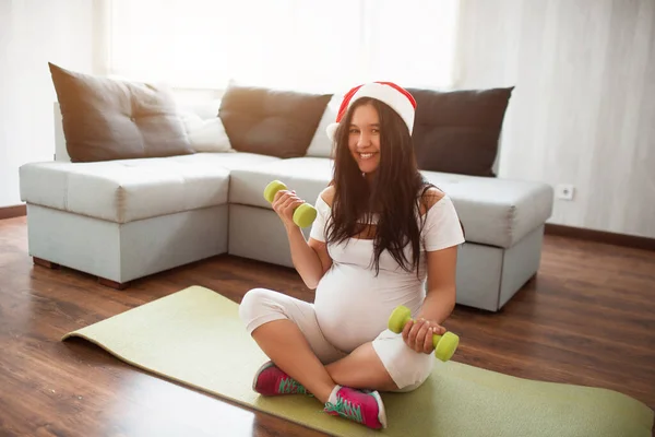 Gott Nytt År och God Jul En gravid kvinna i en röd tomte hatt sitter på en oyga matta och ler mot kameran. — Stockfoto
