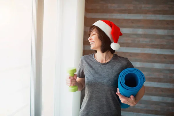 건강 한 성인 여성은 집에서 운동을 한다. 새해나 크리스마스 시간입니다. 잘 지어 진 성인 여성이 일을 마치고 방에서 쉬고 있습니다. 녹색 만두와 파란색 요가 매트를 들고. — 스톡 사진