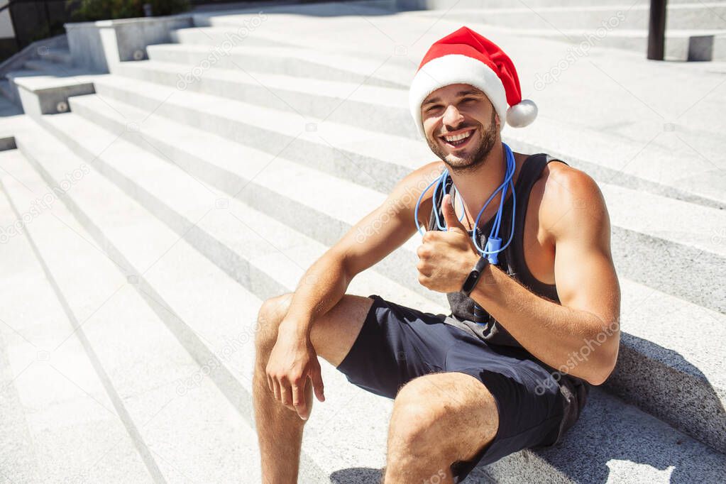 Young man exercising outside. Guy wearing christmas santa hat and show big thumb up. Like symbol. Strong powerful athletist sit alone outisde on steps under the sun.