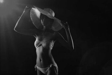 Bikini Yüzücü Kız, Big Straw Beach Hat 'i aksesuarla tut ve kameraya bak. Tek renkli siyah arkaplan, siluet gölge düşük pozlama, yarı vücut portresi