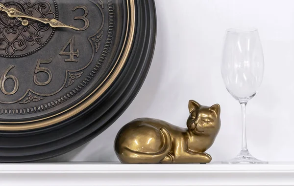 Statuette of a cat against the background of a brown clock and a wine glass.
