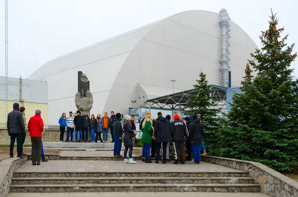 Grupo de turistas no monumento a liquidatários de acidente em Chernobyl NPP no fundo do objeto "Abrigo" sobre a quarta unidade de energia na Zona de Exclusão — Fotografia de Stock