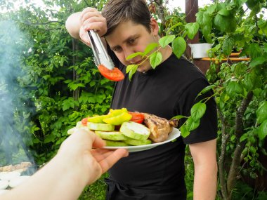 Yakışıklı bir adam kızarmış sebzeleri ızgara maşasıyla tabaktan çıkarır. Doğada eğlence..