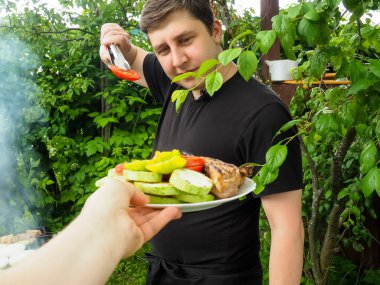 Yakışıklı bir adam kızarmış sebzeleri ızgara maşasıyla tabaktan çıkarır. Doğada eğlence..